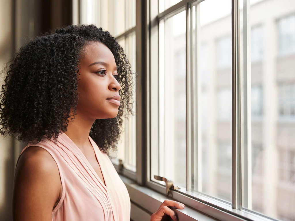 Woman with curly hair wearing a pale pink sleeveless top, looking out a sunlit window with one hand resting on the sill.