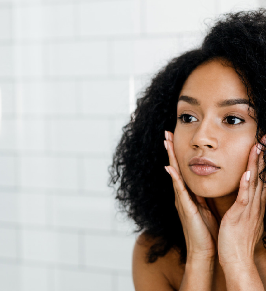 Woman with curly hair gently resting both hands against her cheeks in a white-tiled bathroom.
