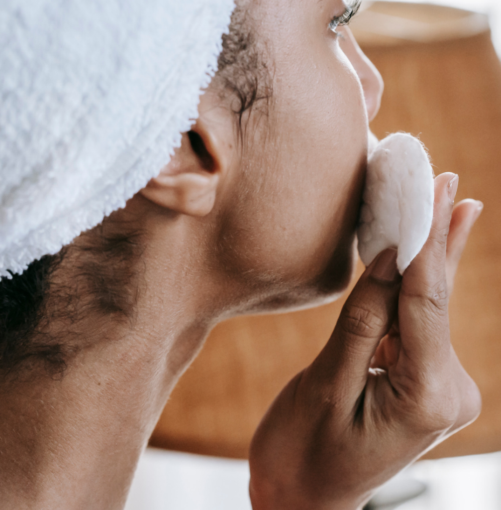 Close-up of a woman with her hair wrapped in a white towel, wiping her cheek with a round cotton pad.