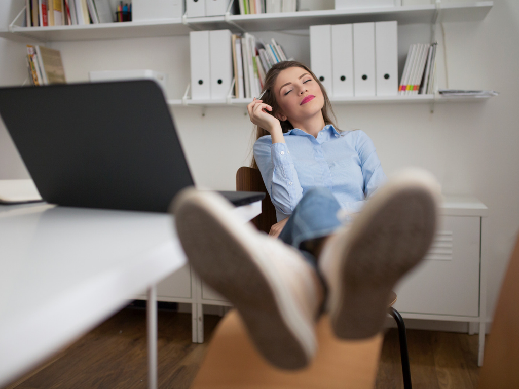 Woman leaning back in an office chair with eyes closed and feet resting on a desk beside an open laptop.