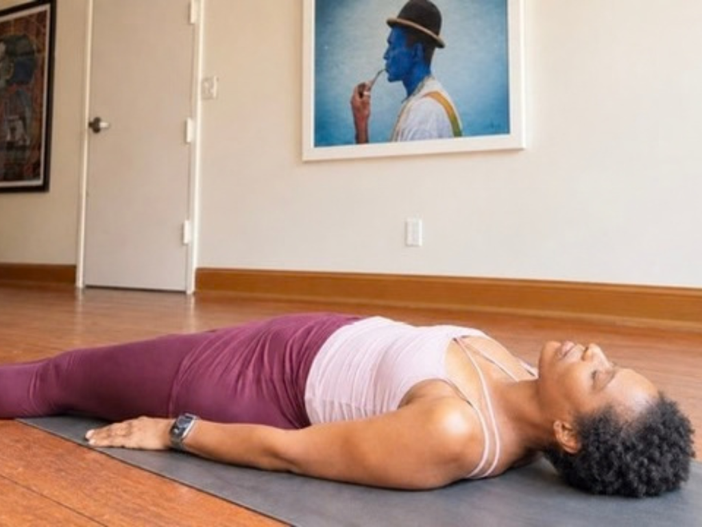 Michelle Wagman resting in Savasana on a yoga mat, eyes closed and arms relaxed beside her, wearing a white top and burgundy pants.