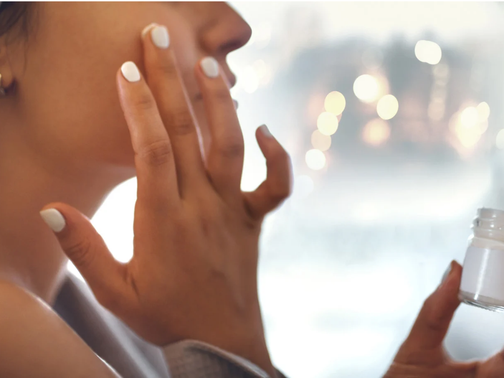 Close-up of a woman applying skincare to her cheek with her fingertips while holding an open jar in her other hand.