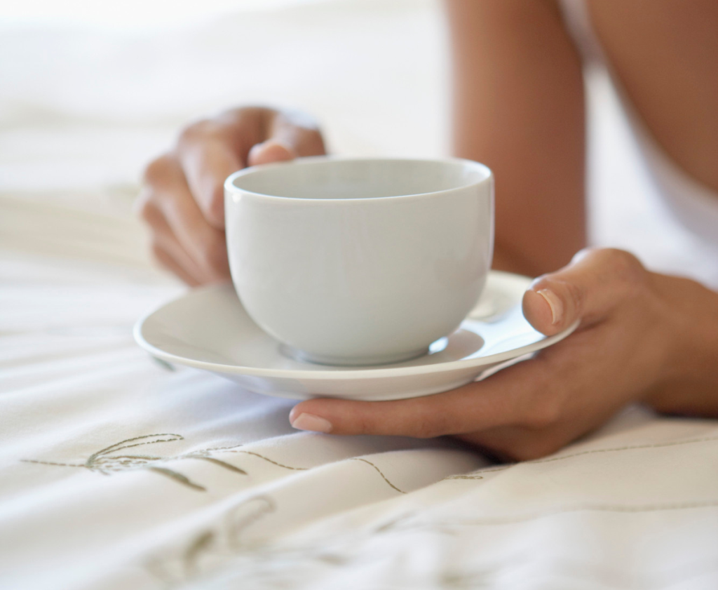 Close-up of a person holding a white cup and saucer over cream-colored bedding.