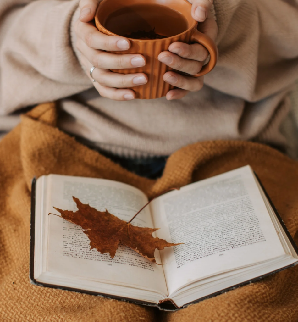 Hands holding a rust-colored mug above an open book with an autumn leaf on its pages, resting on a mustard blanket.