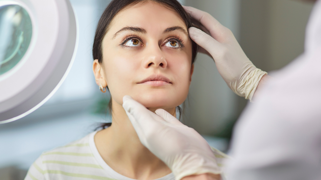 A skincare professional wearing gloves examines a woman’s face beside a magnifying lamp.