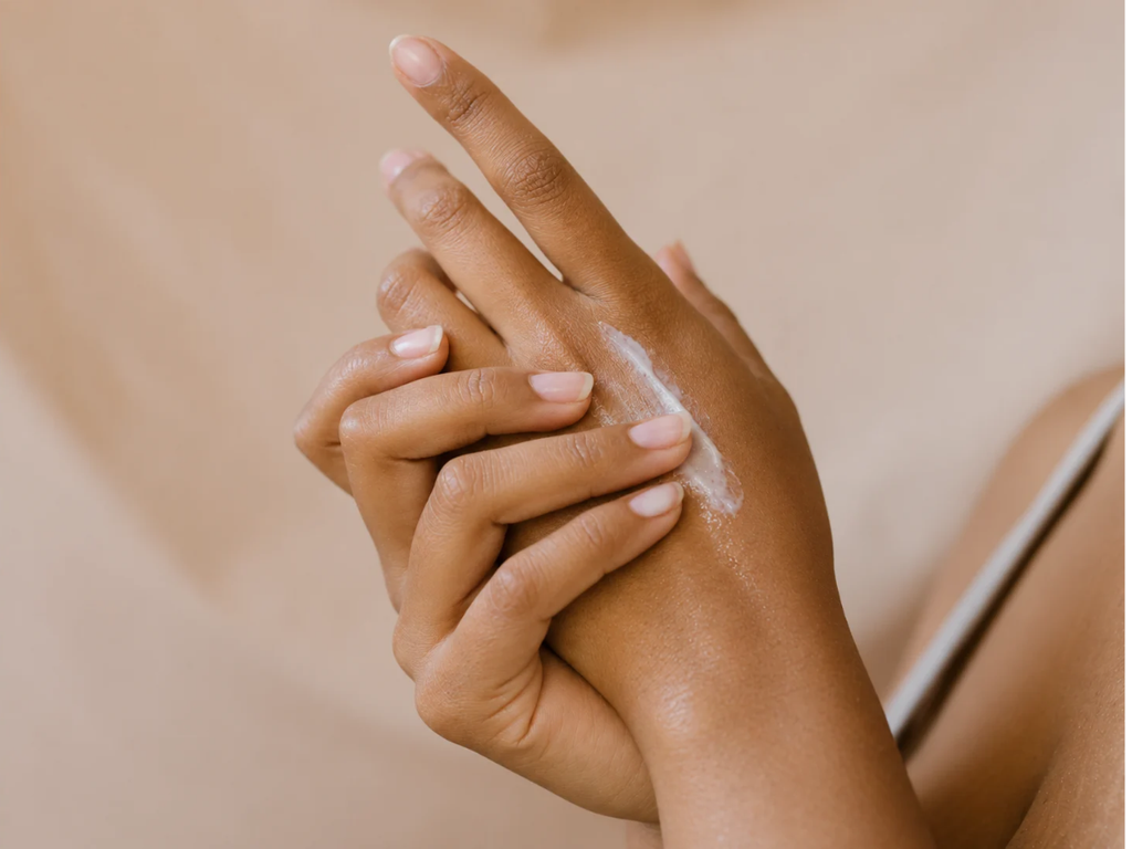 Close-up of brown hands gently massaging white moisturizer into the back of one hand against a warm beige background.