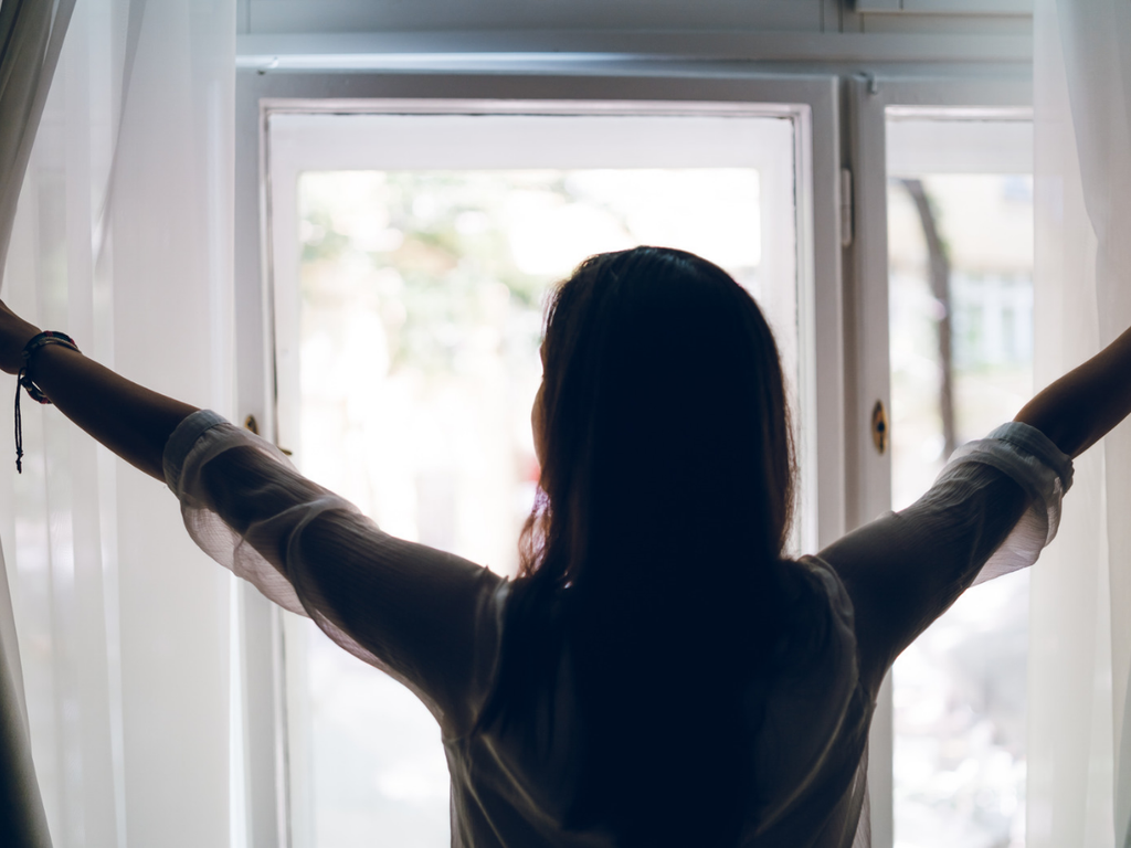 Woman seen from behind opening white curtains to let morning light through a window.